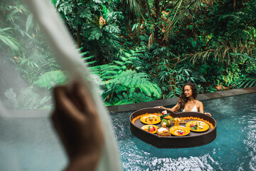 Girl relaxing and eating floating breakfast in jungle pool on luxury villa in Bali. View through window. Valentines day or honeymoon surprise. Tropical travel lifestyle.