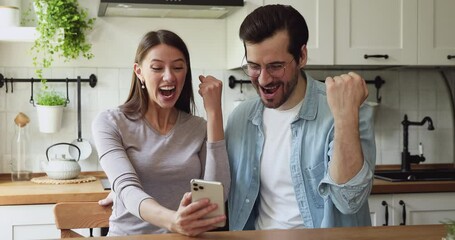 Couple sit at dining table in kitchen check lottery on-line app on smartphone see monetary win feels excited celebrating moment of victory. Sport game fan or online auction gambling winners concept