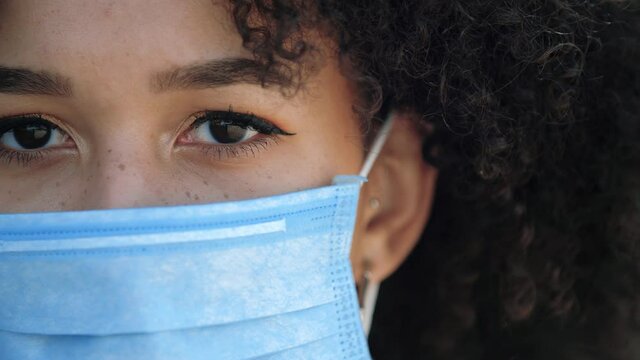 Macro close up beautiful american woman with blue protective mask on face from coronavirus covid outbreak second wave of pandemic looking at camera outdoor street. Brown eyes multiracial ethnic girl