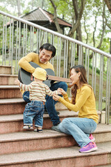 Vietnamese young man playing guitar and singing for his dancing little son, his wife sitting on steps near by