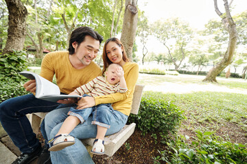 Adorable little kid sitting on laps of his mother and yawning as he is bored of his father reading a book to him