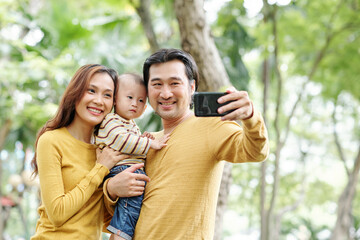 Fototapeta premium Happy positive Asian man taking selfie with his pretty wife and adorable little son