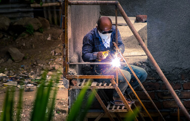 An Indian craftsman welding the iron staircase at the construction site with a cloth face mask due to covid-19 pandemic.