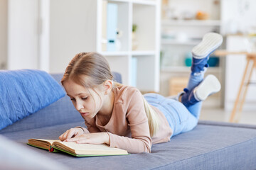 Full length portrait of cute blonde girl reading book or studying while lying on couch in cozy home interior, copy space