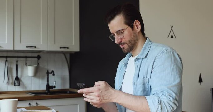 Man in glasses sit in home kitchen hold smartphone enjoy distant informal pleasant chat with friend. Businessman using cell app, check e-mail, surfing web, spend free time have fun on internet concept