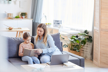 Wide angle portrait of loving adult mother watching cute girl reading book or studying while sitting on couch in cozy home interior, copy space