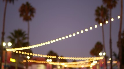 Decorative staring garland lights, palm trees silhouettes, evening sky. Blurred Background. Street decorated with lamps in California. Festive illuminations, beach party, tropical vacations concept.
