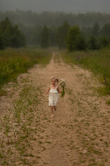 A little girl in a white dress runs along the path with a bouquet of daisies and smiles. Image with selective focus.