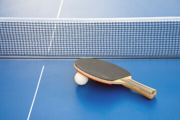 Black wooden racket for ping pong and white ball lying on the blue table next to the net
