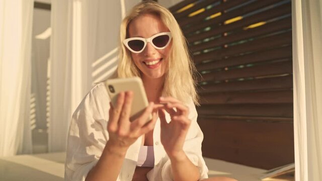 An attractive young blonde woman wearing sunglasses is using her smartphone sitting in the bungalow on the beach in the morning