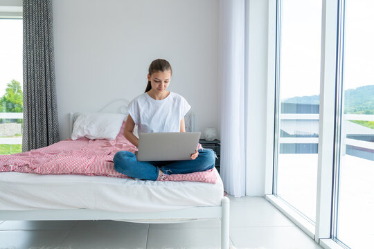 Happy stylish girl with laptop lying on bed in white modern room. Young casual woman shopping online or working online from home