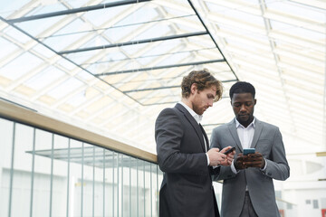 Waist up portrait of two successful businessmen wearing suits using smartphones while standing on glass balcony in contemporary office building under glass roof, copy space