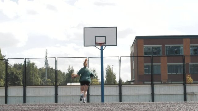 Female basketball player in shorts and white socks on professional court running with ball and successfully thowing ball to the net. Handhelded footage. Outdoors