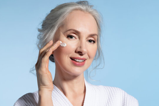 Beautiful senior woman wearing bathrobe applying daily skin care moisturizing nourishing facial cream 