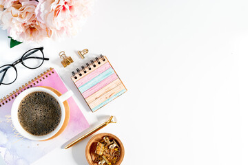 Flat lay of white working place table background with cup of coffee putting on it. Flat lay notebook, glasses, peonies, golden paper binder clips and pen. Desktop mock up planner with copy space