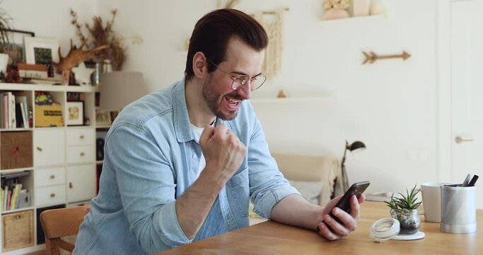 Millennial guy in glasses sitting at table in living room holding smart phone read great news feels very happy. Scream with joy got on-line auction betting results win money. Gambler winner concept