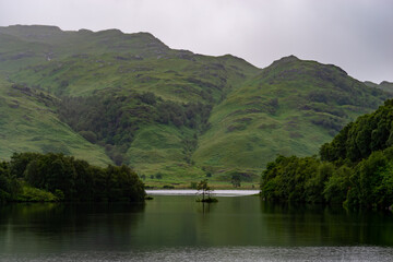 A lonely tree on a lake surrounded by water with mountain ridge building up behind it during a hazy day.