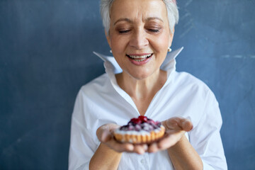 Cropped shot of cheerful attractive grandmother in white shirt holding piece of freshly baked berry pie for birthday, having joyful facial expression, smiling broadly. Bakery and pastry concept