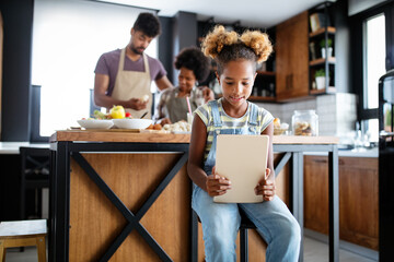 Cute african american girl using a tablet while her parents preparing food in kitchen