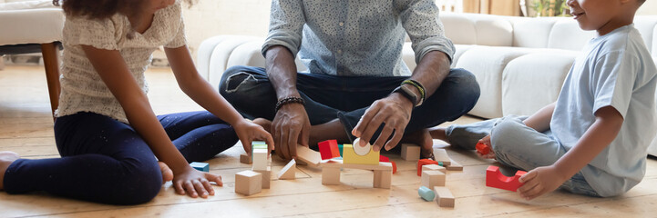 Close up wide banner panorama view of playful african American father with small kids sit on floor play with building bricks, biracial dad with little children construct with wooden blocks at home - Powered by Adobe