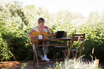 Fototapeta premium Portrait of an elderly woman playing guitar and watching online lessons on laptop while practicing in the garden. Senior woman happy and enjoy life after retired. Online training