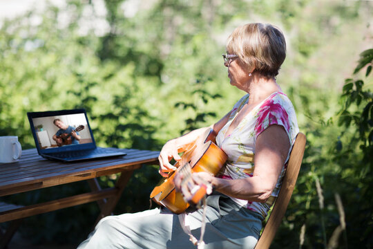 Portrait of an elderly woman playing  guitar and watching  online lessons  on laptop while practicing in the garden. Senior woman happy and enjoy life after retired. Online training