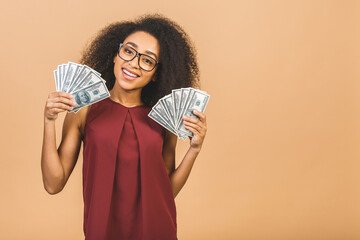 Happy winner. Portrait of african american successful woman 20s with afro hairstyle holding lots of money dollar banknotes isolated over beige background.