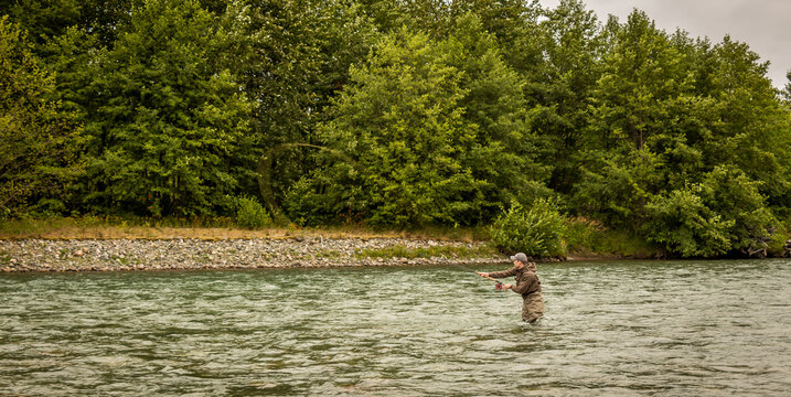 A fly fisherman spey casting while wading in a fast flowing, green, glacial river.