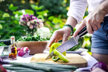 Cutting green peppers by kitchen knife. Woman cooking vegetable salad on table outdoors. Preparation food for garden party