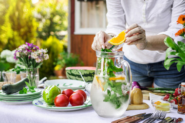 Woman hand putting mint leaf and slice of orange into cold lemonade in carafe. Preparing drink for garden party © encierro