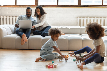 Little biracial brother and sister children sit on warm wooden floor in living room play toys together, happy african American parents couple relax on couch using laptop, ethnic family enjoy weekend