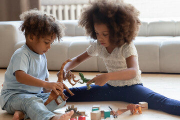 Playful little african American brother and sister kids sit on warm wooden floor at home play rubber dino toys together, cute small biracial children have fun engaged in funny game in living room