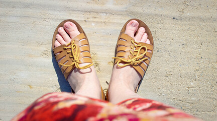 Women's feet in sandals in summer stand on the beach on the sand.