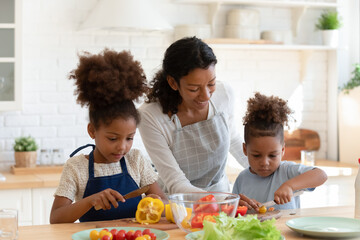 Loving young african American mother or nanny teach little kids prepare healthy food or salad at home kitchen, caring happy biracial mom cooking together with small ethnic children son and daughter