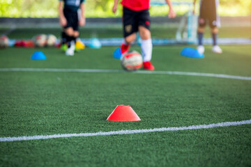 selective focus to red cone marker is soccer training equipment on green artificial turf with blurry kid players training background. Material for trainning class of football academy.