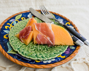 Appetizer of prosciutto wrapped around sweet orange cantaloupe on colorful ceramic dish. 