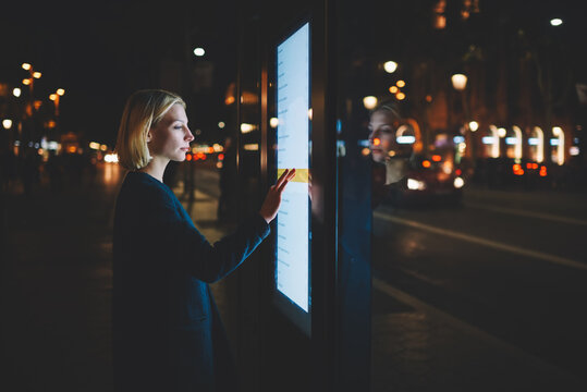 Caucasian female using automated teller machine with big digital screen while standing in night city out-of-focus lights, woman verifies account balance on banking application via modern device icon