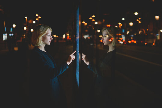 Attractive young woman consults on modern big timetable outdoors, female caucasian tourist touching information kiosk digital screen while standing at night city with beautiful lights on background