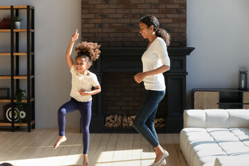 Happy young biracial mom have fun dancing with cute funny little preschooler daughter in modern living room, smiling african american mother or nanny enjoy leisure home weekend, relocation concept