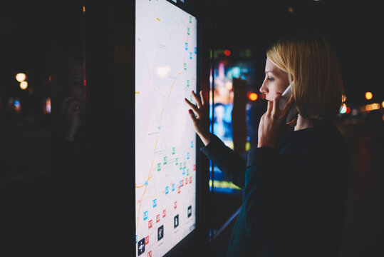 Young woman having a telephone conversation while touching digital display of smart modern bus station, charming female using city computer for touristic information while talking on mobile phone