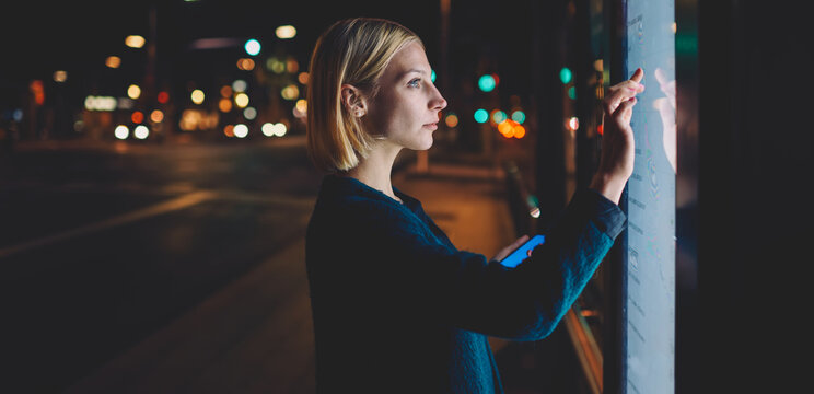 Female caucasian tourist touching information kiosk digital screen while standing outdoors at night city and holding smart phone in the hand, attractive young woman consults on modern big timetable