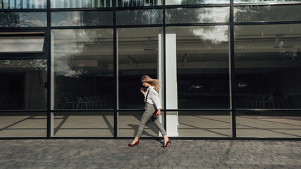 Panoramic full length shot of young business woman goes by. Skyscraper in the center of the city with glass windows. Concentrated girl hurrying to work. Manager, CEO, clerk, boss. Copy space.

