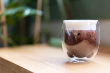 The cold chocolate top is filled with milk foam, put in 2 clear bear shaped face glasses, placed on a wooden table next to the green leafy tree in coffee shop.