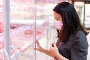 close up young asian woman wear protective mask and standing in front of freezer to decision to choose meat product on aisle shelf  for new normal lifestyle concept