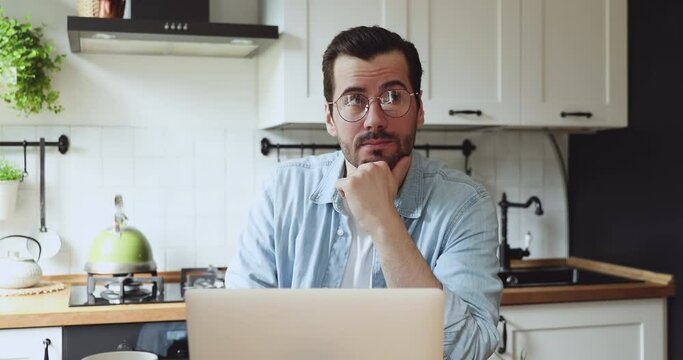 Thoughtful doubtful guy in glasses sit at dining table in home kitchen work on laptop thinking touch chin looking into distance search problem issue solution, ponder question consider new idea concept