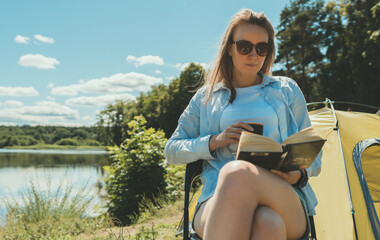 Woman reading a book near camping tent by the lake.