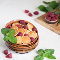 Trendy food pancake cereal, mini pancakes in a bowl with fresh raspberries and mint on a light textile background, selective focus