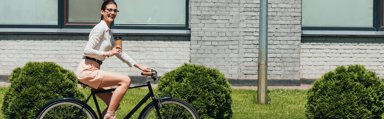 panoramic shot of happy businesswoman in glasses riding bicycle and holding paper cup