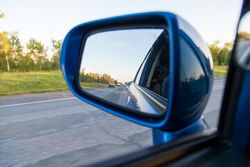 Rear view in the side mirror of a blue sedan with the reflection of heavy traffic on a country asphalt road on a summer day with green trees on the sides of the highway. Traffic Laws.