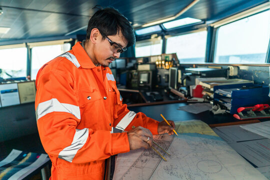 Filipino deck Officer on bridge of vessel or ship wearing coverall during navigaton watch at sea . He is plotting position on chart